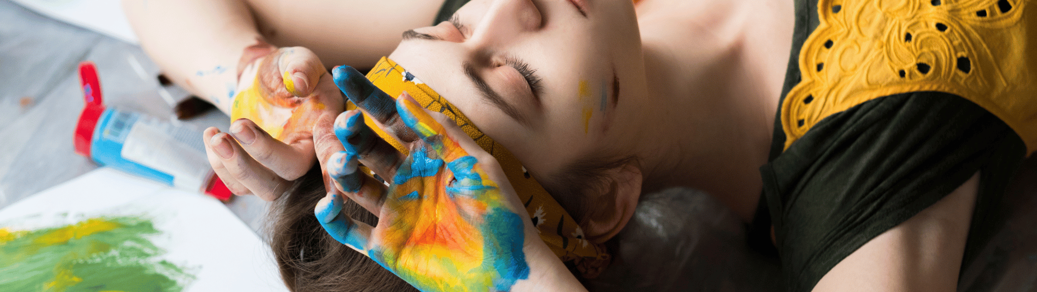 ragazza sdraita con le mani sulla testa e i palmi rivolti verso l'alto sporca di pittura con colori per dipingere sullo sfondo